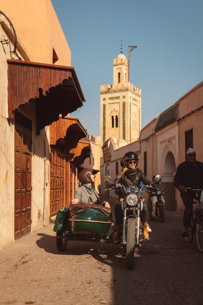 A vintage motorcycle and green sidecar drive through a narrow, sunlit street in a traditional Medina. A woman drives while a passenger sits in the sidecar; both are wearing helmets and sunglasses. A tall, ornate minaret stands in the background against a clear sky, surrounded by classic ochre-colored buildings.