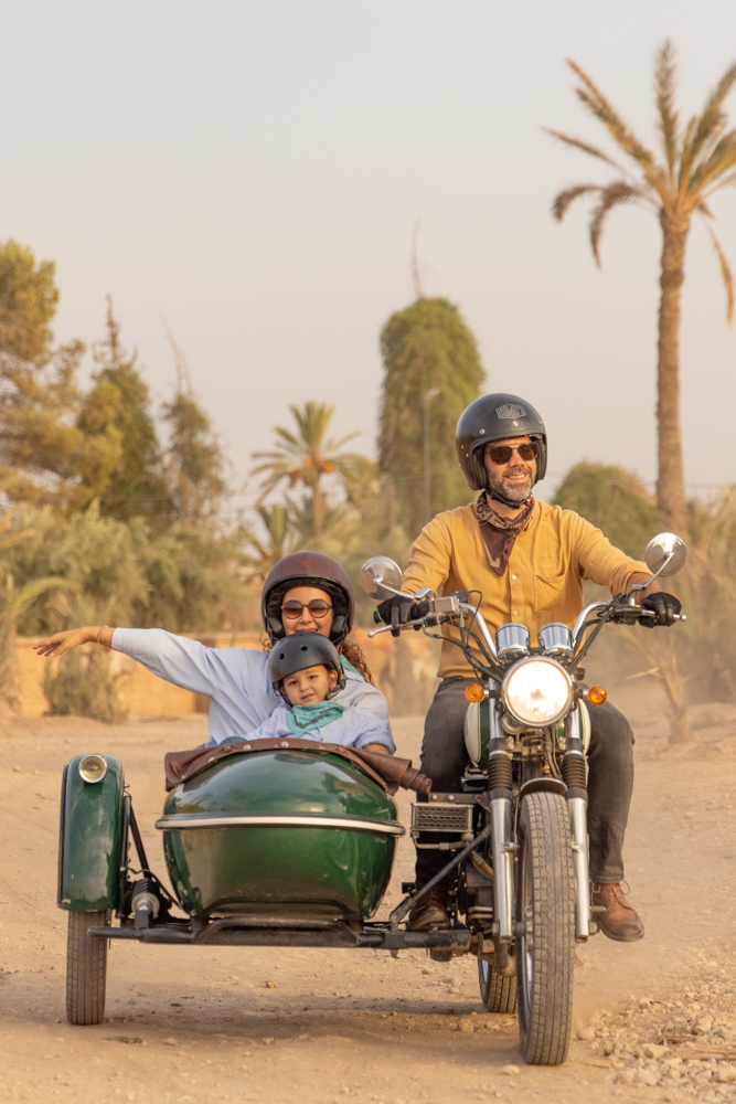 Kids enjoy a ride on a vintage motorcycle with a green sidecar along a dusty path lined with palm trees. All three passengers wear helmets and sunglasses, smiling as they travel through a warm, sunny landscape.