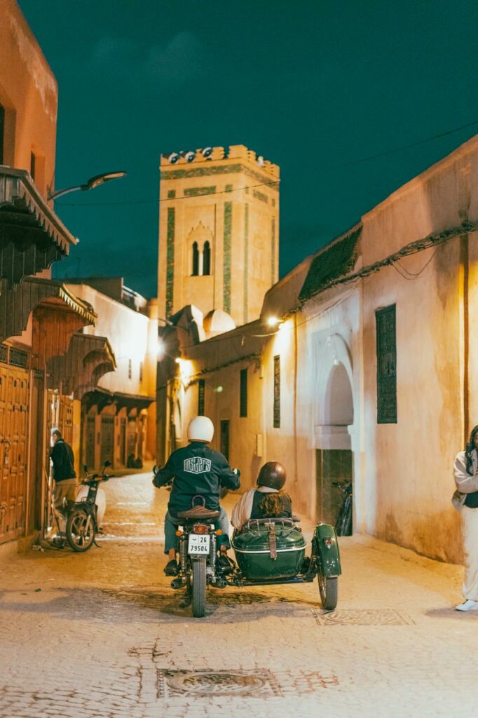 A rear view of a vintage motorcycle and a dark green sidecar as they navigate a narrow, cobblestone alleyway in the heart of the Medina at night. A traditional Moroccan minaret stands tall in the background, beautifully lit against the night sky. The warm, ambient lighting reflects off the ochre walls, capturing a quiet and intimate moment of exploration.